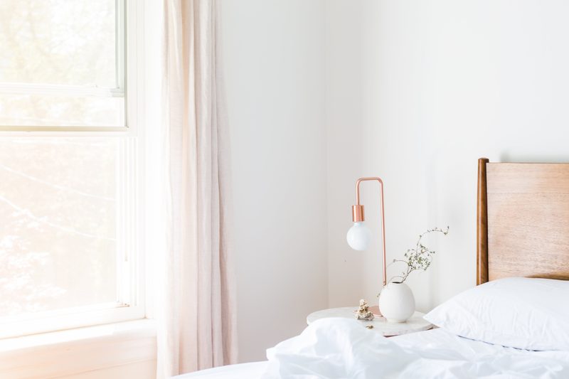 A tidy bedroom with white bedding, a wooden headboard and morning light through a sheer curtain