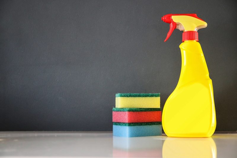 A yellow spray bottle and a stack of three sponges on a white surface against a dark gray wall